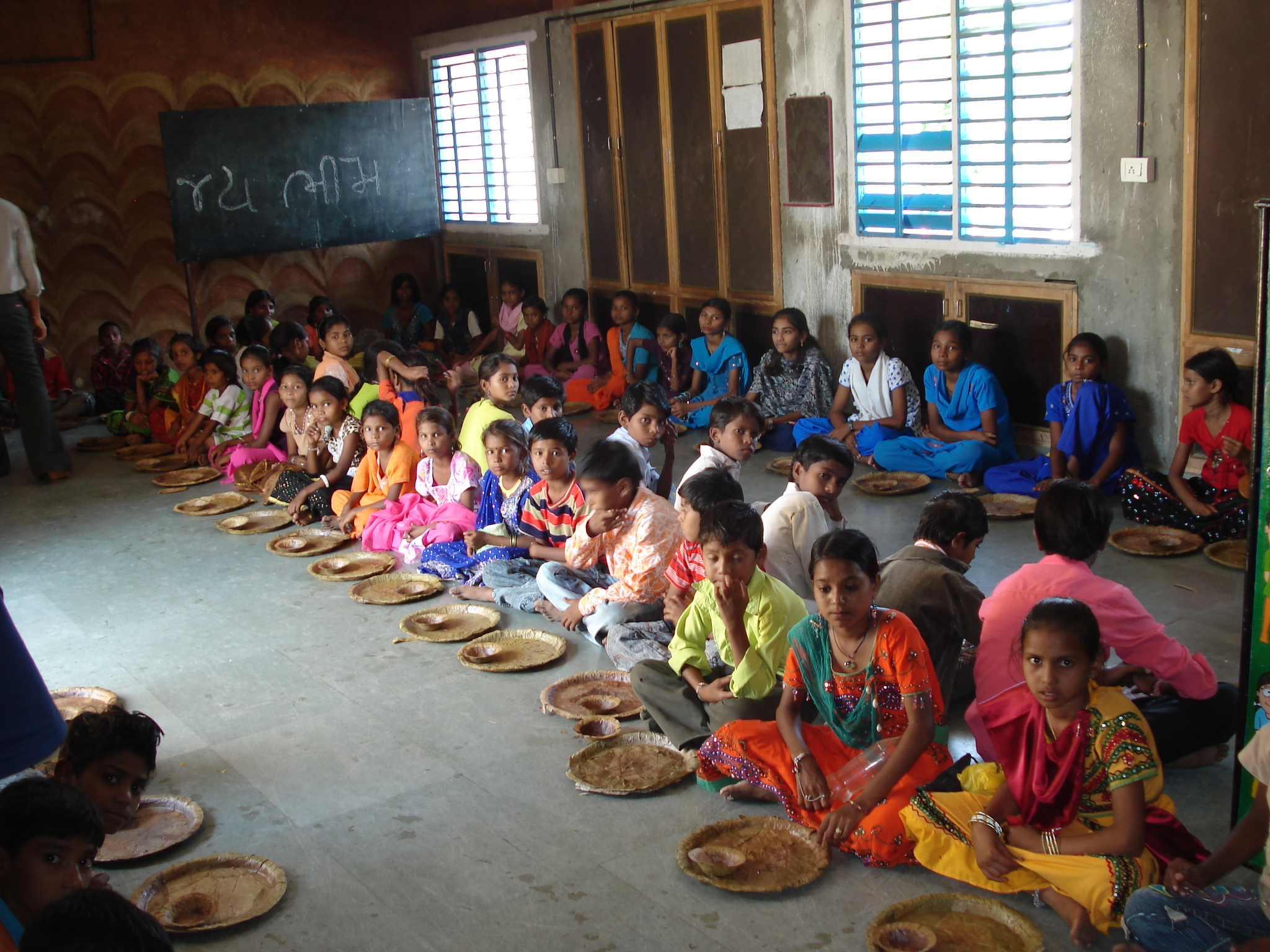 Kanak-kanak menunggu makan tengah hari sekolah di Rayka (juga Raika), sebuah kampung di luar bandar Gujarat. Salam Jai Bhim yang tertulis di papan hitam menghormati ahli undang-undang, reformis sosial, dan pemimpin Dalit B. R. Ambedkar.