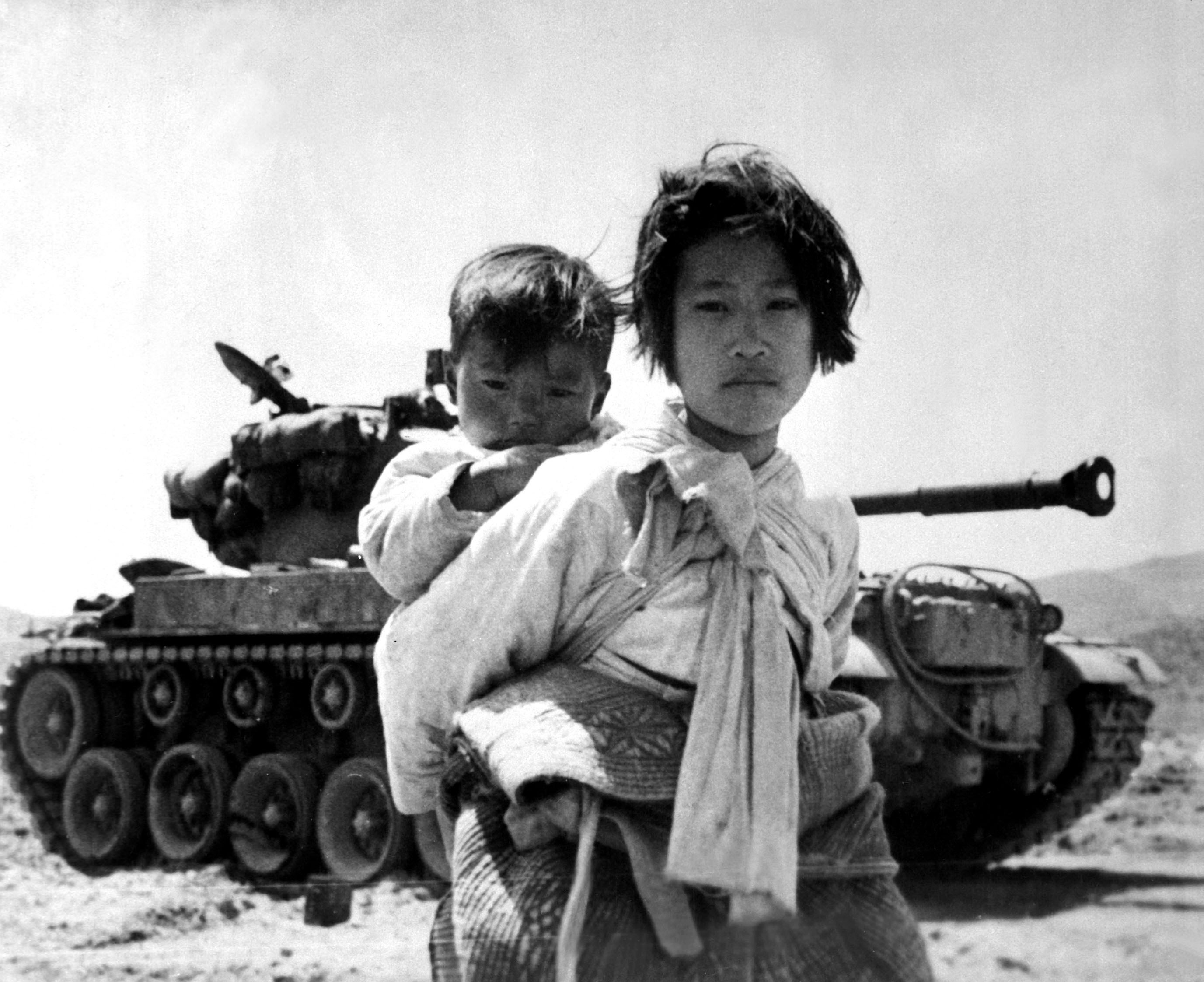 A young girl with her brother on her back, passing by a stalled M-26 tank, June 1951.