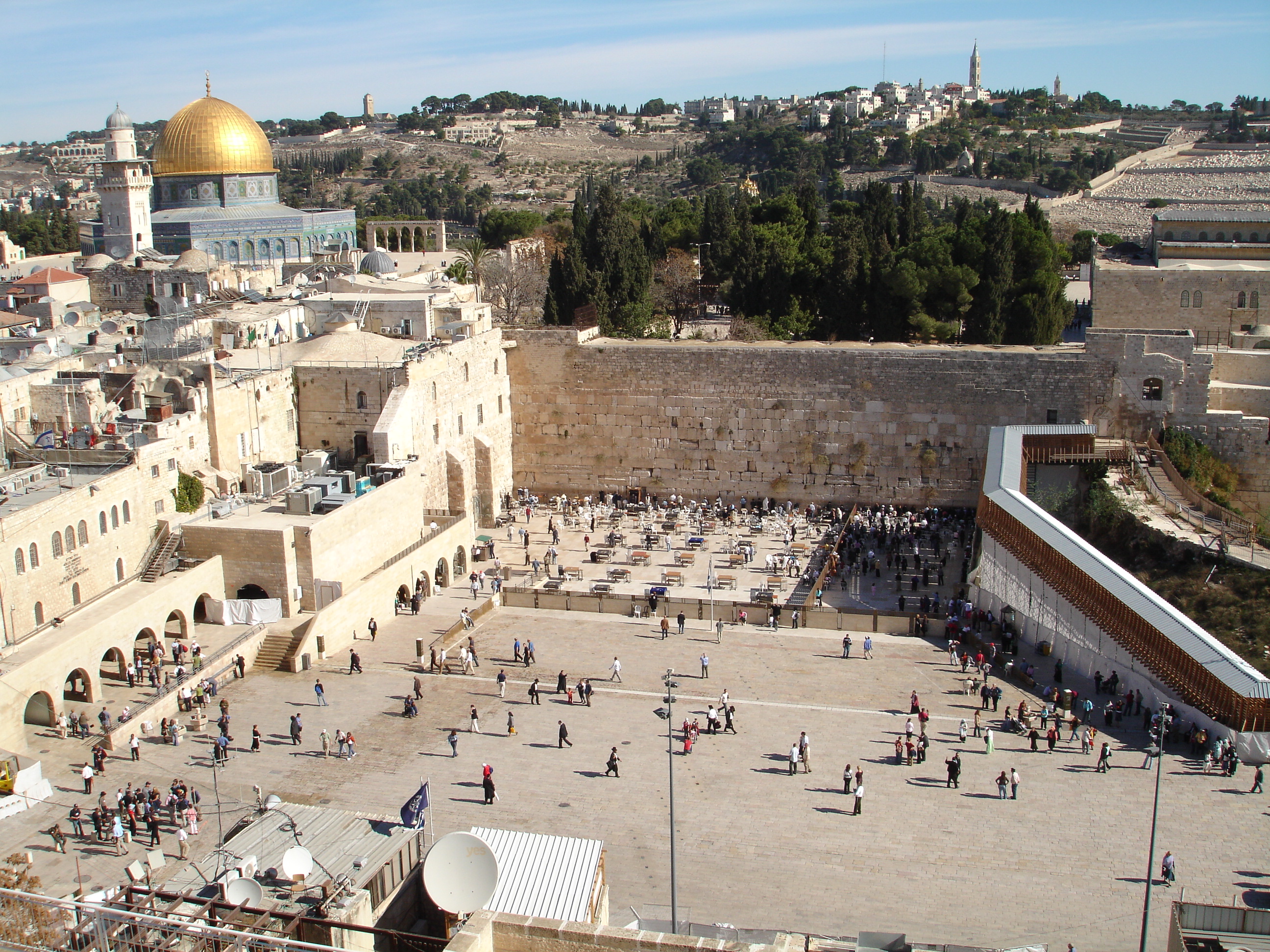 alt=A large open area with people bounded by old stone walls. To the left is a mosque with large golden dome.