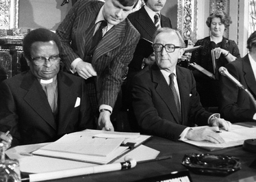Uskup Abel Muzorewa menandatangani Perjanjian Lancaster House duduk di sebelah Menteri Luar Negeri Inggris Lord Carrington.