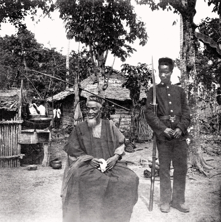 Pemimpin Temne, Bai Bureh dilihat di sini pada tahun 1898 selepas penyerahan dirinya, duduk santai dengan pakaian tradisionalnya sambil memegang sapu tangan, manakala seorang askar Pasukan Sempadan Afrika Barat Sierra Leone berkawal di sebelahnya.