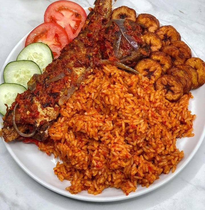 Nasi Jollof dengan ikan goreng dan plantain, dihiasi dengan timun dan tomato